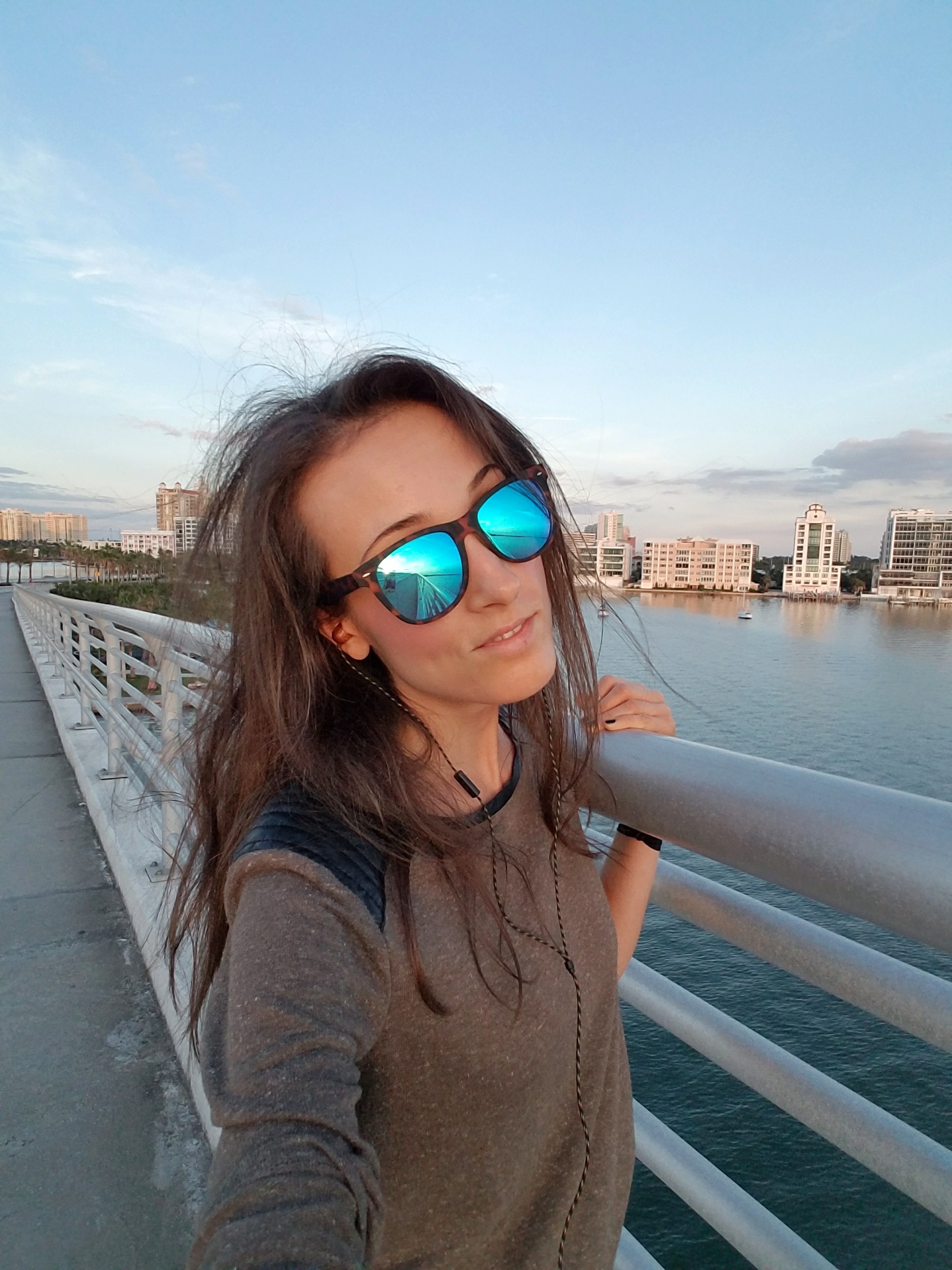 Woman wearing sunglasses with earbuds, walking on a bridge with a water view