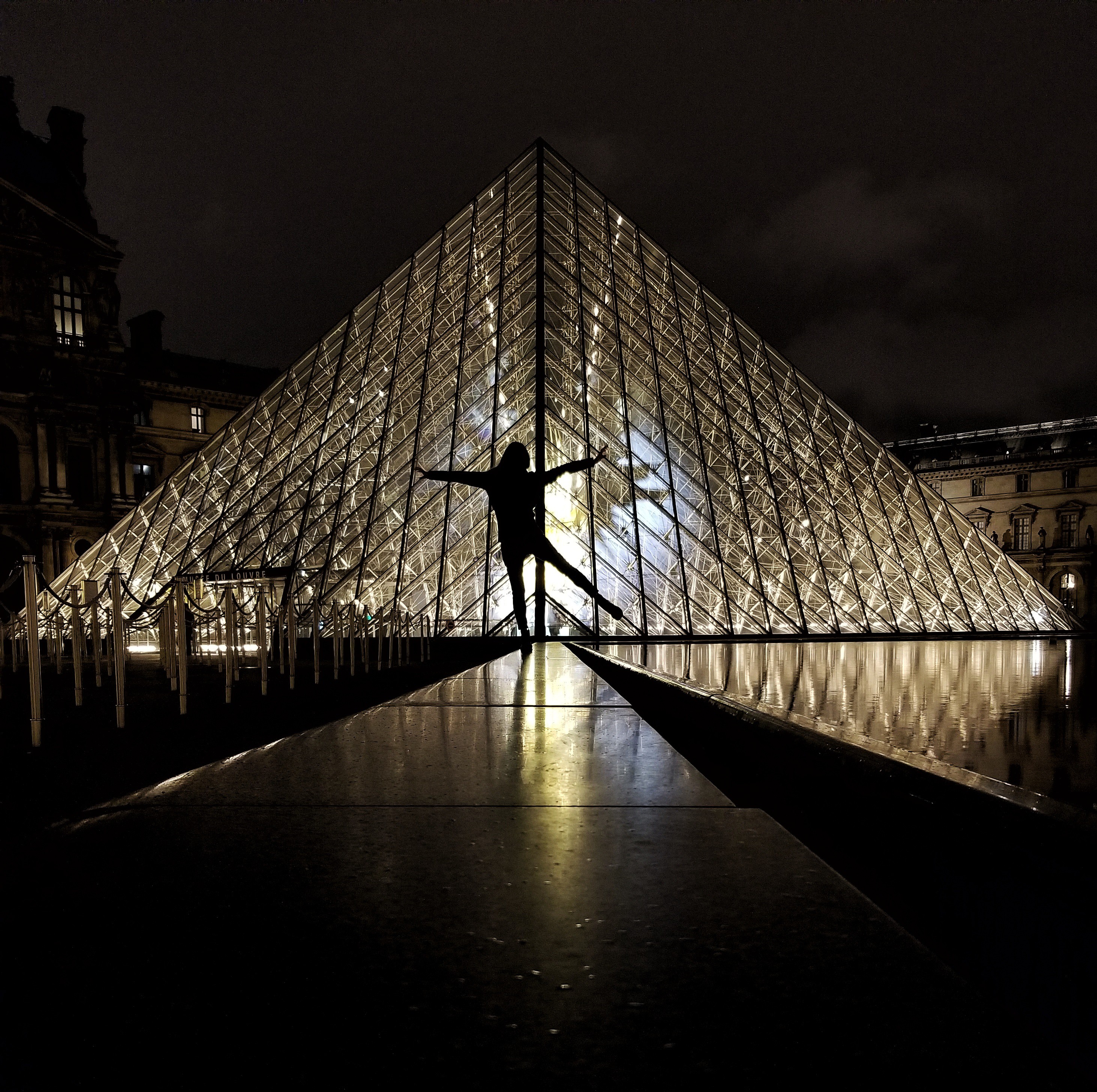 Silhouette of a woman standing in front of the Louvre Pyramid at night