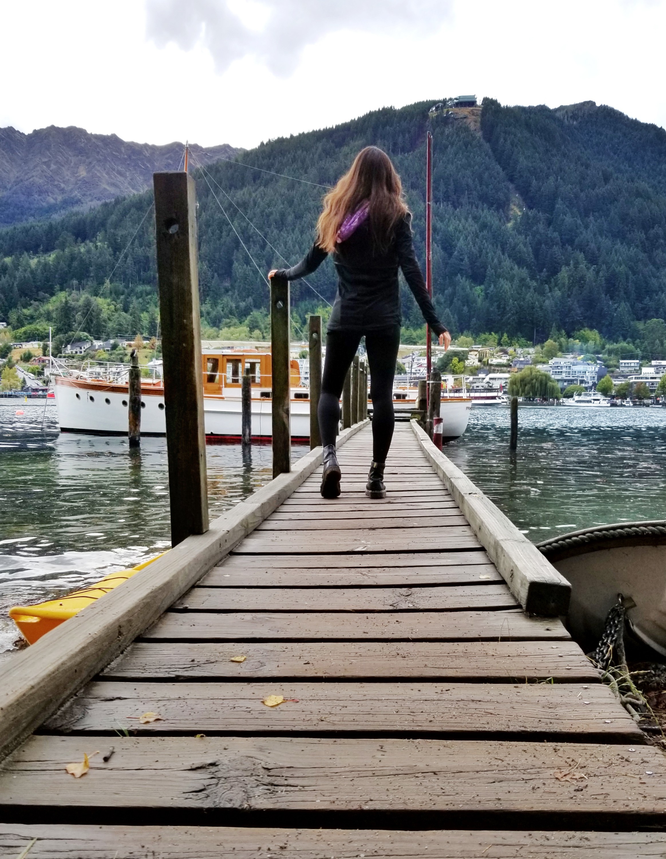Woman walking on a dock with a boat at the end and mountains in the distance