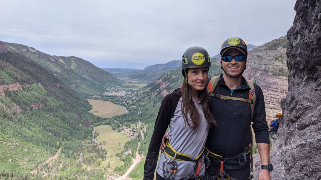 Woman and man with helmets and climbing gear, standing on the edge of a cliff face
