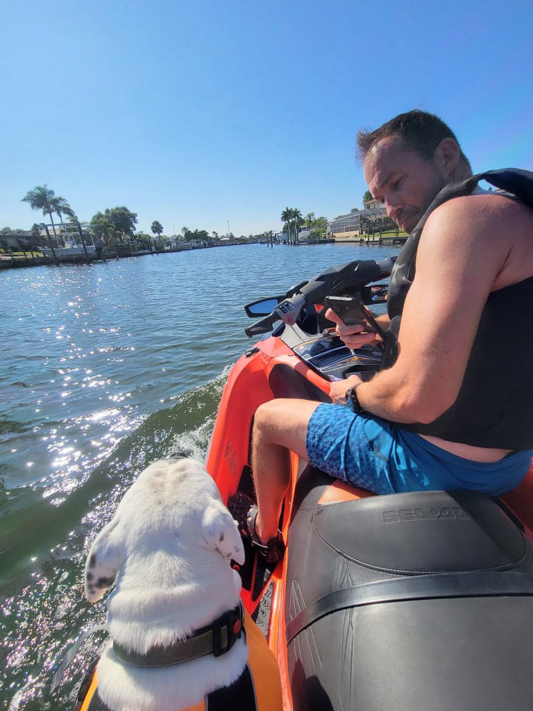 Man in a life jacket sitting on a jet ski with a white dog in a life jacket 