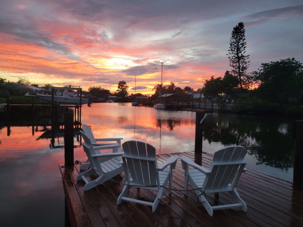 A dock with four white Adirondack chairs at sunset over a canal