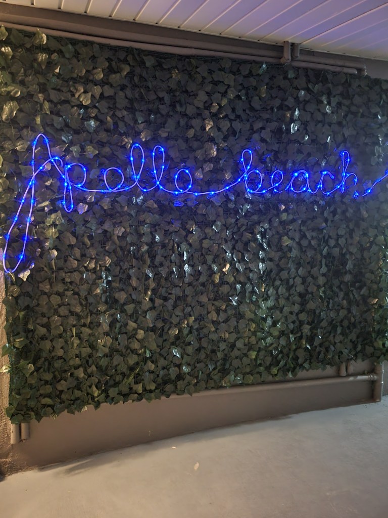Wall of fake leaves with the words "Apollo Beach" lit up at night