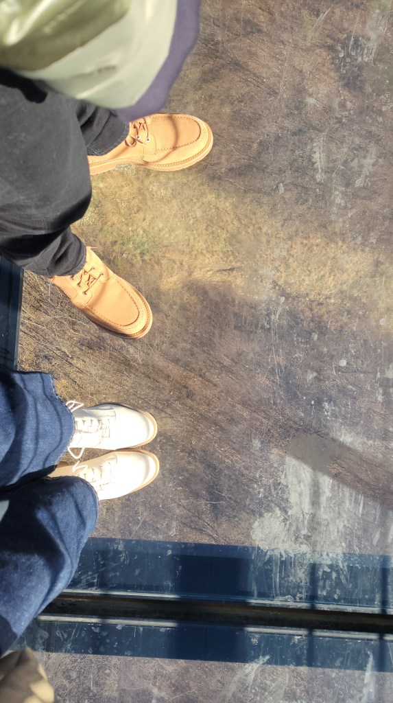 Photo looking down at the boots of two people standing on a glass-bottom bridge