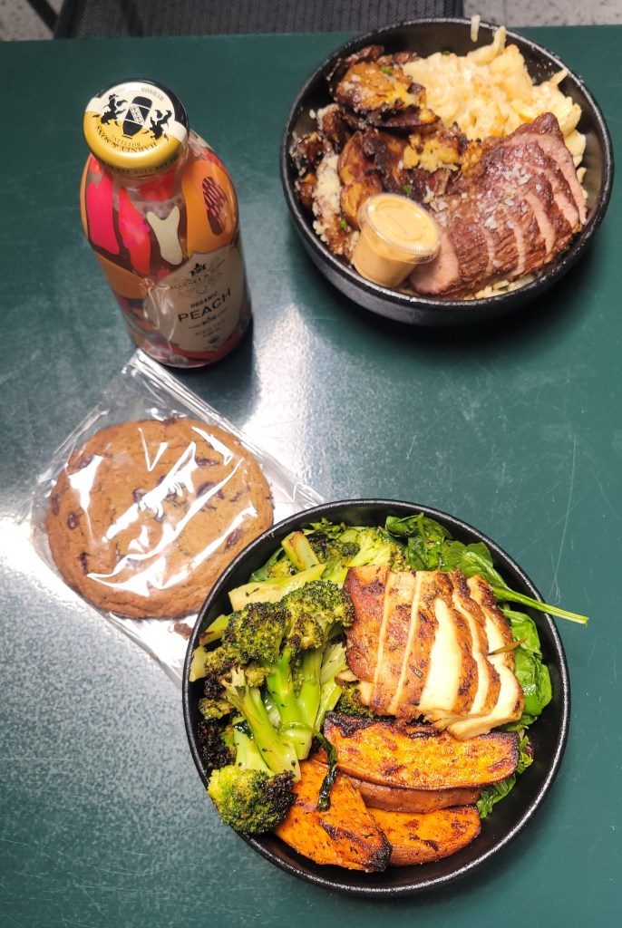Two bowls of food on a green table with a bottle of tea and chocolate chip cookie